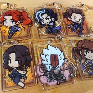 May include: Six acrylic keyrings featuring cartoon characters with gold-coloured key rings. Each keyring has a different character design, with vibrant colours and detailed artwork. The keyrings are rectangular with a brown border.