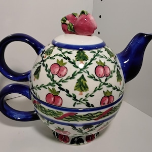 Puede incluir: Tetera redonda de cerámica con asa y boquilla azules. La tetera está decorada con un patrón de frutos rojos, hojas verdes y pequeños árboles verdes sobre fondo blanco. La tapa está rematada con dos frutos rojos.