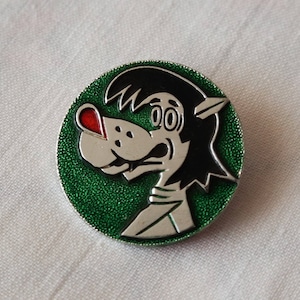 May include: A green and silver pin with a cartoon wolf wearing a white shirt. The wolf has a red nose and black hair.