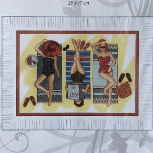 Può includere: Un kit per punto croce contato con una scena di spiaggia con tre figure che prendono il sole. Il kit include il testo "Crystal Art" e "Vacanza al mare". Il disegno finito misura 25 x 17 cm.
