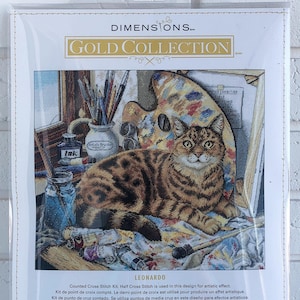 Puede incluir: Un kit de punto de cruz contado que presenta un gato sentado en un lienzo salpicado de pintura con pinceles, una paleta y tubos de pintura. El kit se titula "Leonardo" e incluye las dimensiones 14 pulgadas por 12 pulgadas (35,5 cm x 30,4 cm).