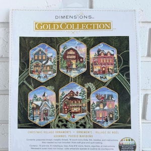 May include: A Dimensions Gold Collection cross stitch kit for six Christmas village ornaments. The package features six hexagonal designs of winter buildings, including a bakery and toy shop. It includes thread, 18 count ivory Aida, and needles.