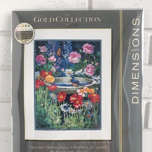May include: A counted cross stitch kit featuring a colorful garden scene with a bluebird perched on a birdbath. The kit includes a variety of flowers, including red poppies, pink roses, and white daisies. The design is titled "Garden Reflections" and is by Arleta Pech. The kit measures 11 inches by 15 inches.