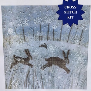Könnte beinhalten: Kreuzstich-Set mit einer verschneiten Winterlandschaft mit zwei braunen Kaninchen, die durch ein Feld mit weißen Blumen laufen. Der Himmel ist blau mit fallenden weißen Schneeflocken. Der Text "CROSS STITCH KIT" befindet sich in einem blauen Sternenausbruch.