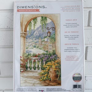 May include: A Dimensions Needlecrafts cross stitch kit titled "Terrace Arch." The kit features a detailed cross-stitch design of a scenic terrace with flowers and a view of a coastal town. The package measures 9 x 16 inches (22.8 cm x 40.6 cm).