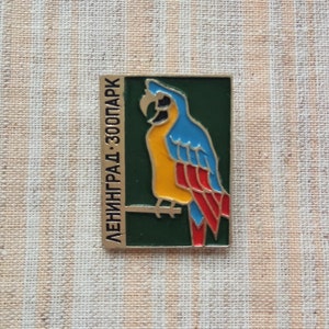 May include: A vintage enamel pin featuring a colorful parrot on a green background. The text "ЛЕНИНГРАД - ЗООПАРК" is written vertically on the left side of the pin.