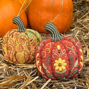 May include: Two decorative pumpkin ornaments with gold hanging strings rest on hay, in front of three orange pumpkins. One ornament is red with yellow floral embroidery and green beads. The other is yellow with red and green embroidery.