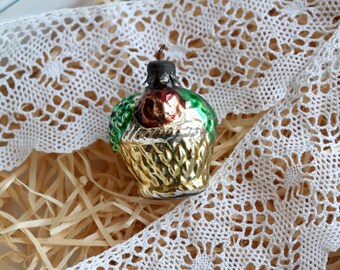 May include: A vintage glass Christmas ornament shaped like a basket filled with fruit. The ornament is decorated with gold, green, and red paint. It is sitting on a bed of shredded paper and a white lace doily.