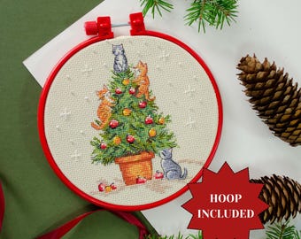 May include: A cross-stitch embroidery hoop featuring a Christmas tree decorated with cats. The tree is in a terracotta pot and adorned with red ornaments. The hoop is red, and the text "HOOP INCLUDED" is on a red starburst.