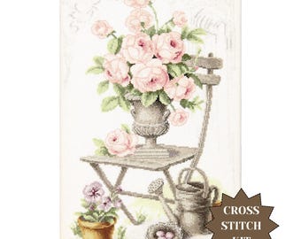 May include: Cross stitch kit featuring a floral still life. A vase of pink roses sits on a small wooden table, with a watering can and a pot of purple flowers. The words "Cross Stitch Kit" are in a starburst.