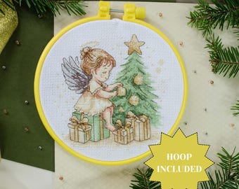 May include: Cross-stitch embroidery kit featuring a Christmas angel decorating a tree, set in a yellow hoop. The angel is depicted with wings and a halo, surrounded by wrapped gifts. The text "HOOP INCLUDED" is in a yellow starburst.