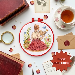 以下が含まれることがあります： 赤いフレームのクロスステッチ刺繍枠には、本を読む女性が描かれ、花と蝶に囲まれています。「HOOP INCLUDED」の文字が赤い星型でデザインされています。本、お茶、虫眼鏡などのアイテムも含まれています。