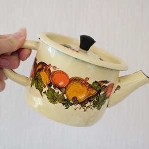 Könnte beinhalten: Eine weiße Emaille-Teekanne mit einem schwarzen Knopf auf dem Deckel. Die Teekanne hat ein Fruchtdesign mit Orangen, Zitronen und grünen Blättern.