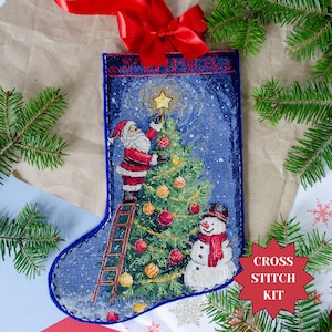 May include: A blue Christmas stocking cross stitch kit with a festive design. It depicts Santa Claus placing a star on a Christmas tree, with a snowman nearby. The kit includes a red bow and the text "Cross Stitch Kit".