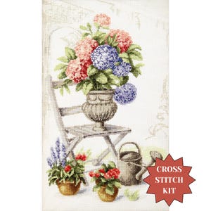 May include: Cross stitch kit featuring a floral still life. The design includes a wooden chair, a large vase with pink and blue hydrangeas, and potted flowers. A watering can is also present. The words "Cross Stitch Kit" are in a red starburst.