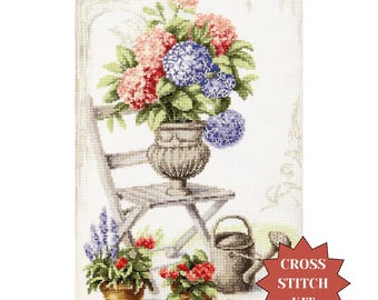 May include: Cross stitch kit featuring a floral still life. The design includes a wooden chair, a large vase with pink and blue hydrangeas, and potted flowers. A watering can is also present. The words "Cross Stitch Kit" are in a red starburst.