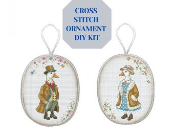 May include: Two oval cross-stitch ornaments featuring anthropomorphic geese in vintage clothing. One goose wears a brown coat and hat, the other a blue dress and hat. The text "CROSS STITCH ORNAMENT DIY KIT" is above the ornaments.