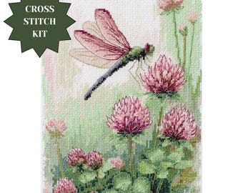 May include: A cross stitch kit featuring a detailed embroidered design of a dragonfly with translucent pink wings hovering above a cluster of vibrant pink clover flowers and green leaves. The text CROSS STITCH KIT is printed in white on a dark green star.