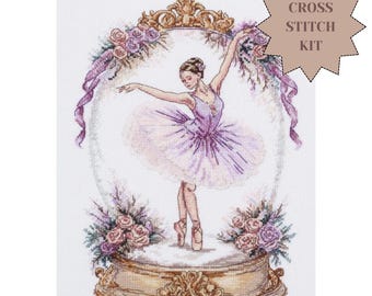 May include: A cross stitch kit featuring a ballerina in a purple tutu dancing inside an ornate gold frame. The design includes pink and purple floral arrangements at the base and top of the frame, with ribbons on the sides. The text CROSS STITCH KIT is visible in the upper right corner.