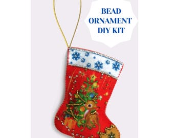 May include: A red Christmas stocking bead ornament DIY kit. The stocking features a deer and Christmas tree design with gold and silver beads. The top of the stocking has a white border with blue snowflakes. A gold string is attached for hanging.
