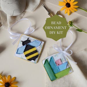 May include: A bead ornament DIY kit featuring two square fabric ornaments with white ribbon loops. One ornament displays a black and yellow bee with light blue wings, while the other shows a geometric floral pattern in green, blue, and pink.