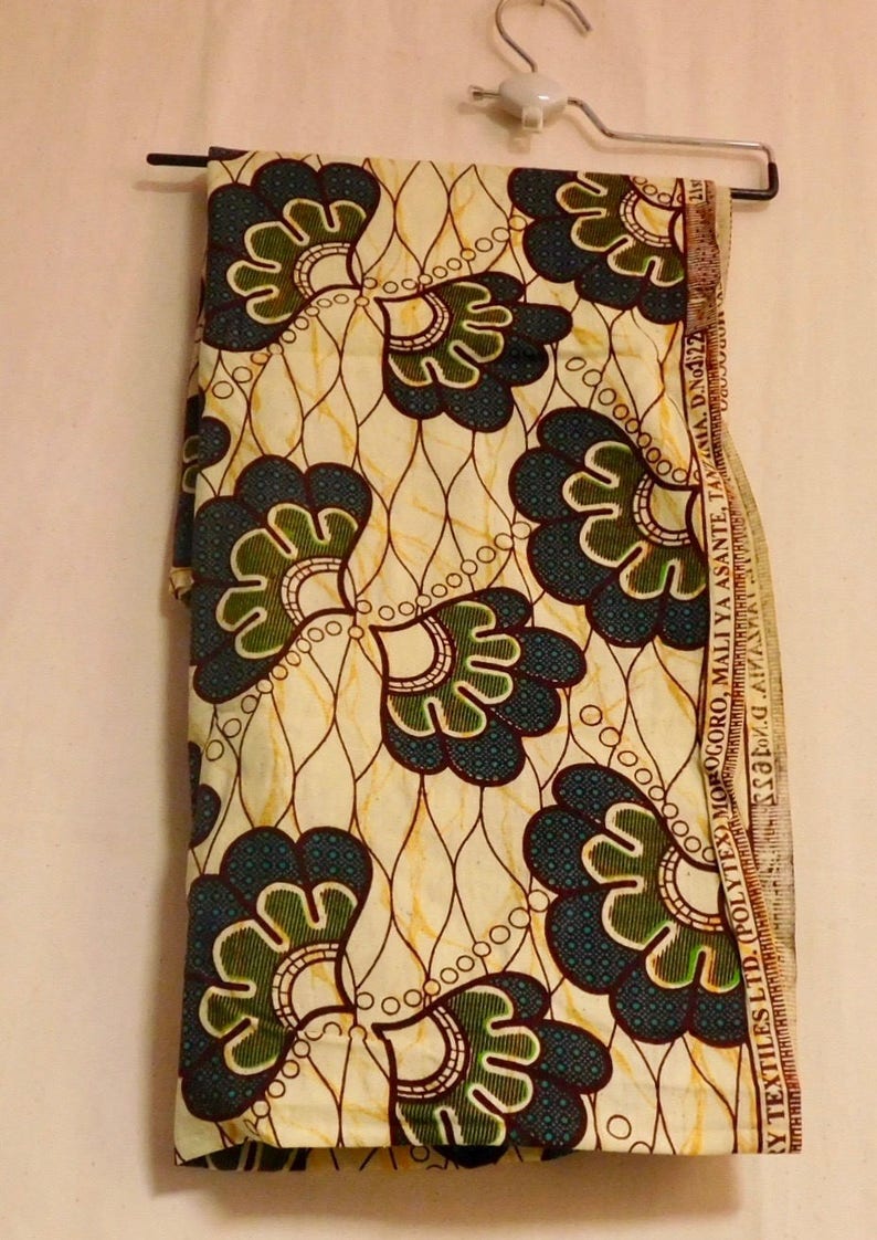 May include: A cream-colored fabric with a repeating pattern of green, blue, and brown stylized flowers. The fabric has a label that reads "(Y TEXTILES LTD. (POLYTEX) MOROGORO, MALI YA ASANTE, TANZIMA. D.No1/22".