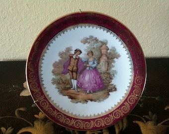 May include: A decorative porcelain plate with a gold rim and a burgundy border. The plate features a painted scene of a couple in a garden setting.