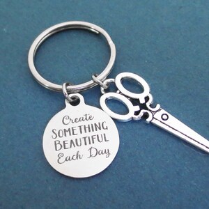 Może przedstawiać: Srebrny brelok do kluczy z okrągłym wisiorkiem z napisem "Tworz coś pięknego każdego dnia" i małą srebrną parą nożyczek.