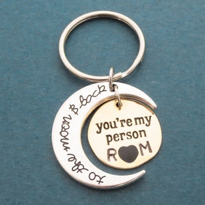 May include: Silver key chain with a crescent moon charm that says "To the moon & back". The charm has a smaller gold circle charm that says "You're my person" with a black heart and the letters "ROM".