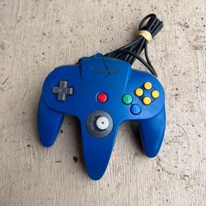 May include: A blue Nintendo 64 video game controller featuring a central analogue stick, a grey directional pad, a red start button, and four yellow C-buttons alongside green A and blue B buttons. A black wired cable is coiled and secured with a rubber band.