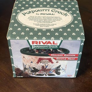 May include: A square cardboard box for a Rival Potpourri Crock electric simmering cooker. The box is dark green with a white clover pattern and features text describing the product, along with an image of a holiday-themed stoneware crock.