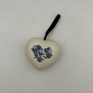 May include: A heart-shaped ceramic ornament in an off-white colour featuring a blue floral pattern painted on the front. The ornament includes a black ribbon loop attached to the top for hanging, providing a simple and functional design element.