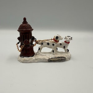 May include: A decorative figurine featuring a dark brown fire hydrant connected by gold-colored chains to two white Dalmatian dogs with black spots and red collars, all mounted on a textured white base resembling a snowy or icy surface.
