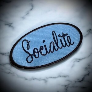 May include: A light blue oval-shaped patch with black stitching around the edges. The word "Socialite" is embroidered in black cursive lettering on the patch.