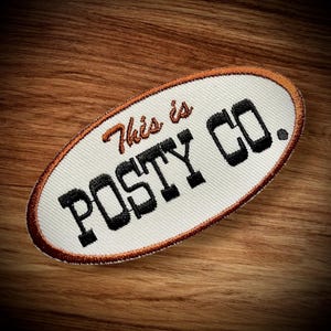May include: An oval-shaped embroidered patch with the text "This is POSTY CO." in black and orange. The patch has a white background with an orange border and is set against a wooden surface.