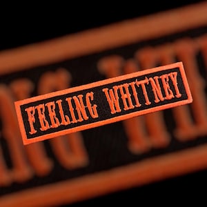 May include: A rectangular patch with the words "FEELING WHITNEY" embroidered in orange thread on a black background. The patch has an orange border and is likely designed to be sewn onto clothing or accessories.
