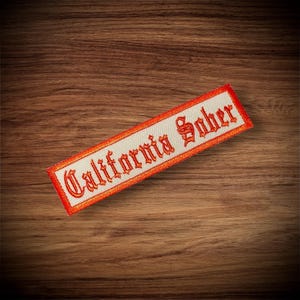 May include: A white fabric patch with an orange embroidered border and the text "California Sober" in orange.