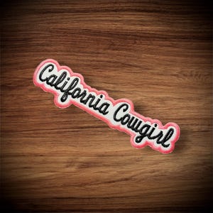 May include: A white embroidered patch with the words "California Cowgirl" in black script lettering. The patch has a pink border and is set against a wooden background.