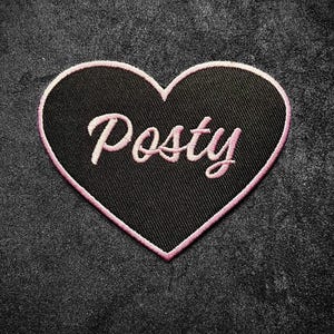 May include: A black heart-shaped patch with a pink embroidered border. The word "Posty" is embroidered in pink cursive script across the center of the patch. The patch is set against a dark gray textured background.