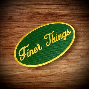 May include: A green oval-shaped patch with a yellow border. The text "Finer Things" is embroidered in yellow on the patch.