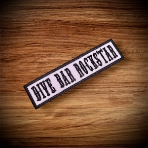 May include: White patch with black embroidered text that reads "DIY Bar Rockstar" on a wood background.