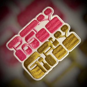 May include: Embroidered patch with the word "GET" in a stylized design. The patch features pink and gold thread on a white background, with a white border. The design is geometric and modern, suitable for clothing or accessories.