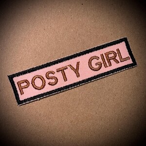 May include: A pink fabric patch with black stitching around the edges. The text "POSTY GIRL" is embroidered in gold thread.