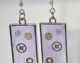 May include: A pair of silver earrings with a rectangular purple background and silver gears. The earrings are hanging from silver hooks.