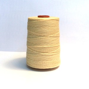 Puede incluir: Una bobina de hilo de algodón de color natural enrollada alrededor de un carrete cónico. El hilo es liso y está enrollado apretado, con un ligero brillo.