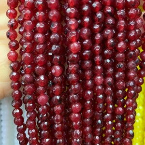 May include: A strand of red faceted gemstone beads. The beads are small and round, with a slightly irregular shape. They have a glossy finish and are strung on a clear thread.