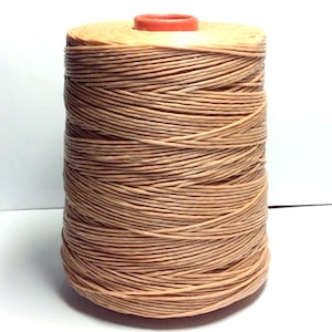 Puede incluir: Una bobina de hilo de lino encerado beige. El hilo está enrollado firmemente alrededor de la bobina, que tiene una tapa de plástico roja.