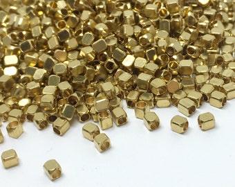 May include: A large pile of gold-colored metal beads, with a few beads scattered on a white surface. The beads are small and have a square shape with rounded edges.