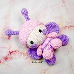 May include: A handmade crochet butterfly toy in shades of pink, purple, and cream. The toy features a rounded head with antennae, large wings, and a striped body. The text "Kasha Craft" is visible on the bottom.