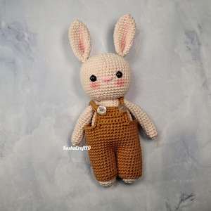 May include: A handmade crochet bunny doll with a light beige body, pink-lined ears, and rosy cheeks. The bunny wears brown overalls with button closures. The doll is approximately 8 inches tall, perfect for a child's toy or nursery decor.